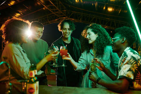 Five happy youthful intercultural people in stylish casualwear clinking with cocktails while...
