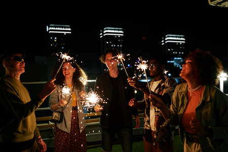 Group of happy youthful multicultural friends with sparkling bengal lights enjoying party in rooftop...