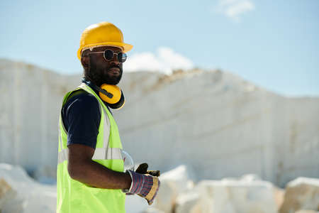 Young confident African American male engineer in workwear, gloves, eyeglasses and protective helmet looking at you during workの写真素材