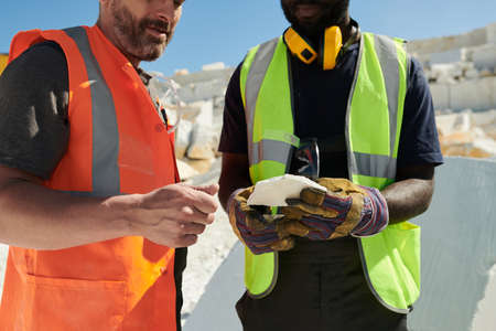 Mid section of two intercultural workers of marble quarry in uniform discussing sort of solid rock...
