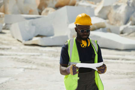 Young African American engineer in gloves, hardhat, uniform and eyeglassess holding paper with...
