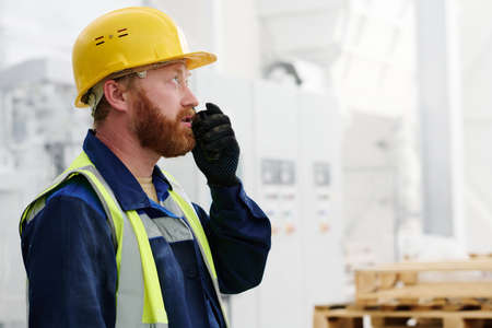 Bearded male worker in uniform, hardhat and black gloves using walkie-talkie while reporting about...