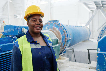 Happy young African American female engineer in hardhat and unidorm standing in workshop with industrial production equipmentの写真素材
