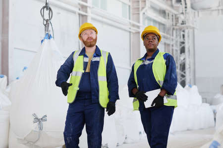 Two young confident colleagues in uniform and safety helmets having discussion of new industrial...