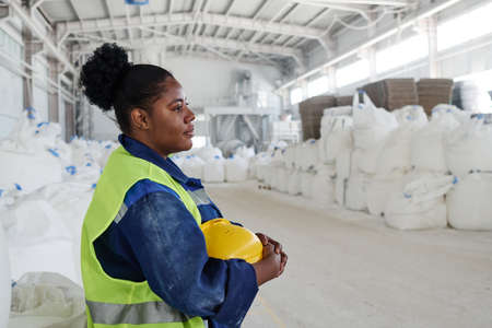 Side view of young black woman in workwear holding yellow safety helmet while standing in front of...