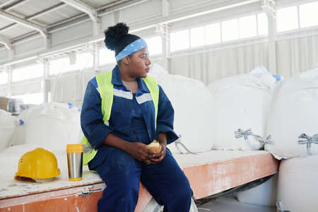 Young tired female worker of warehouse sitting among huge white sacks with loose raw materials and having sandwich and tea for lunchの写真素材