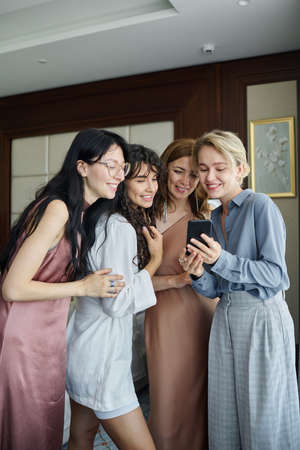 Young blond woman with smartphone showing new photos to her friends in elegant dresses during preparation of bride for weddingの写真素材
