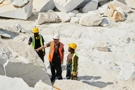 Group of intercultural workers of quarry checking quality of marble rocks while standing in front of one of them on territory of factoryの写真素材