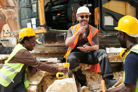 Group of happy middle aged intercultural workers of marble quarry having lunch and communicating while sitting by excavatorの写真素材
