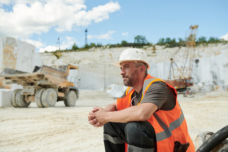 Tired worker of marble quarry sitting in front of camera by workplace against dump truck and working bulldozer with craneの写真素材