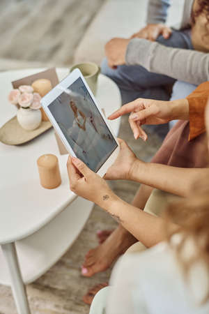 Hand of young woman pointing at wedding dress on screen of tablet held by her girlfriend while...