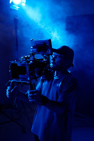 Serious young cameraman with steadicam standing against spotlight in studio lit by blue light and...