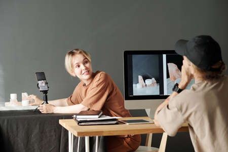 Happy young assistant looking at photographer sitting in front of computer and consulting with him...