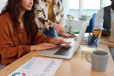 Young businesswoman with pencil pointing at screen of laptop of colleague sitting by desk and correcting electronic documentの写真素材