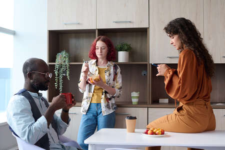 Three young restful intercultural employees in smart casualwear having food, tea and coffee and...