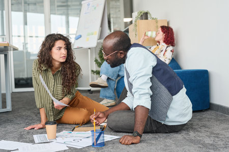 Young confident businesswoman showing financial document to African American male coworker while...