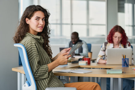 Young successful businesswoman with tablet sitting by workplace in office among her colleagues and...