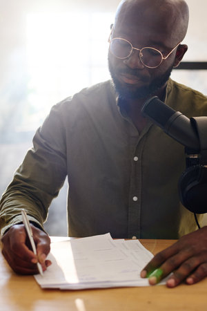 Young bald black man reading questions on paper prepared for interview with guest while sitting by desk in studio in front of microphoneの写真素材