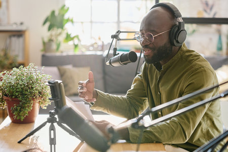 Black guy in headphones talking in microphone while reading plan of discussion and recording new video for online audienceの写真素材