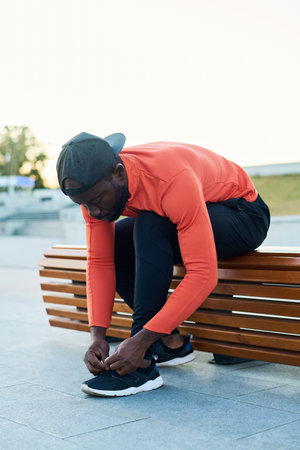 Young sportsman in activewear tying shoelaces of sneakers while sitting on wooden bench in park or urban environmentの写真素材