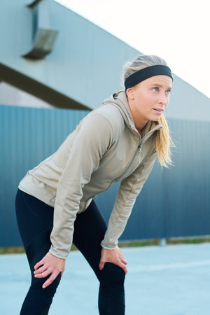 Young woman with legs bent in knees standing in front of camera in urban environment after jogging...