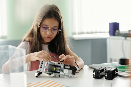 Cute serious schoolgirl creating robot or model of machine during lesson while sitting by desk and connecting parts of new itemの写真素材