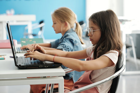 Clever youthful schoolgirl and her classmate typing on laptop keypads while sitting by desk and working with online informationの写真素材