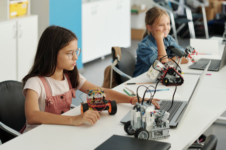 Clever youthful schoolgirl entering information in electronic document and looking at laptop screen while describing toy robotの写真素材