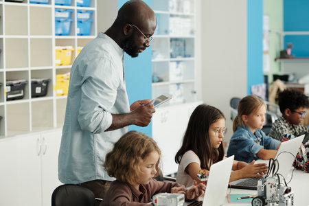 African American male teacher of robotics checking task of schoolchildren sitting in row in front of...
