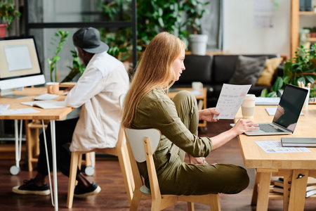 Young businesswoman with financial document looking at laptop screen while sitting by workplace and analyzing online informationの写真素材