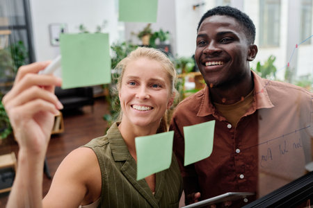 Young smiling blond female employee explaining her colleague working points written on notepapers on...