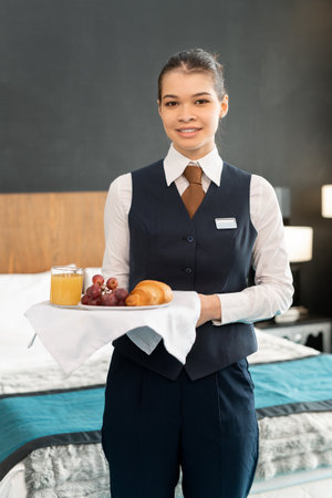 Photo realistic digital collage of happy young elegant female worker of hotel holding tray with breakfast while standing in front of cameraの素材