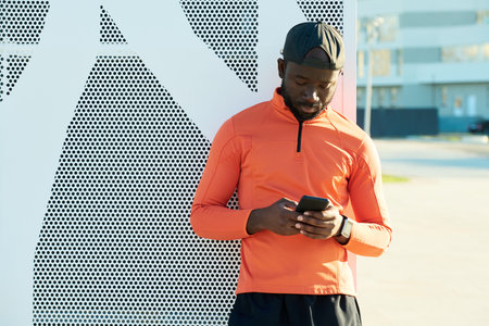 Young sportsman with smartphone texting or looking through list of contacts while standing by wall...