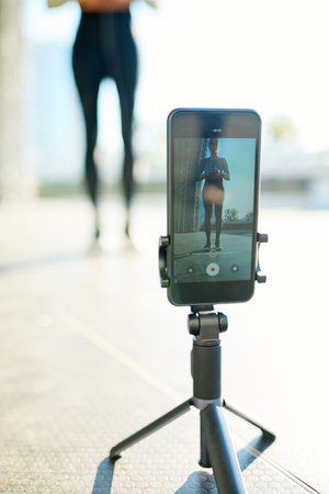 Tripod with smartphone during video record of sportswoman having outdoor workout while standing in urban environmentの写真素材