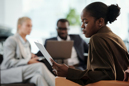 Young modern office worker reading business contract against two intercultural colleagues having discussion at meetingの写真素材