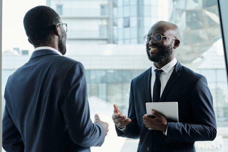 Happy African American businessman with tablet talking to young male colleague in formalwear at working meeting in officeの写真素材