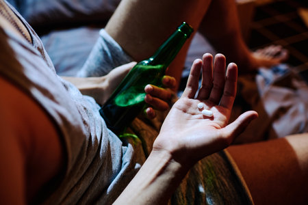 Above angle of hand of young depressed man holding sleeping pills or antidepressants and bottle of alcohol while sitting on bedの写真素材