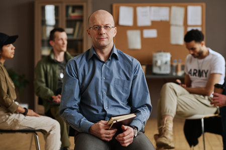 Confident mature male counselor or psychologist with notebook looking at camera while sitting against group of young intercultural patientsの写真素材