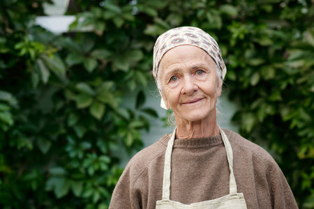Medium shot of smiling senior woman in apron, pullover and headcloth looking at camera while standing against green foliageの写真素材