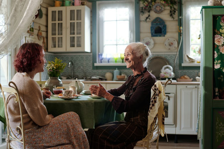 Cheerful senior woman in smart casualwear chatting with her granddaughter by breakfast while sitting...