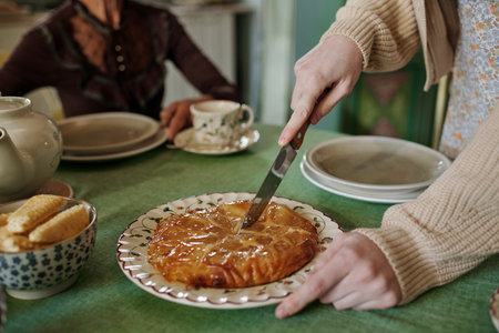 Hands of young woman with sharp knife cutting homemade sweet pie while standing by served table where her grandmother sittingの写真素材