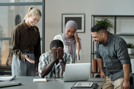 Group of young multicultural students laughing at black man hiding his face while crying by workplace surrounded by cruel classmatesの写真素材