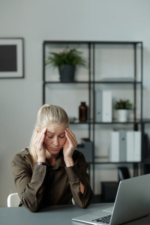 Young tired blond student or businesswoman touching head while sitting by desk in front of laptop...