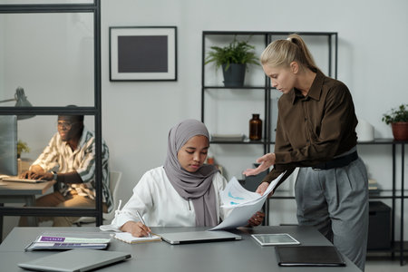 Young blond angry female boss scolding her subordinate in hijab while showing her documents and explaining what to doの写真素材