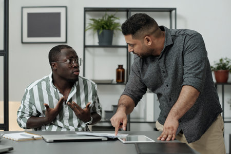 Middle aged boss bending over workplace of African American male subordinate while rebuking him for not carrying out working taskの写真素材