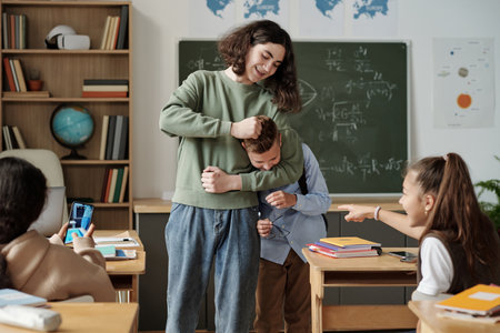 Two girls laughing at junior school learner while one of them making video of their classmate fighting with him in classroomの写真素材