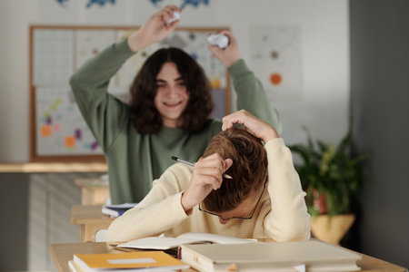 Youthful schoolboy bending over desk and covering his head by hands while his classmate throwing crumpled paper at him at lessonの写真素材