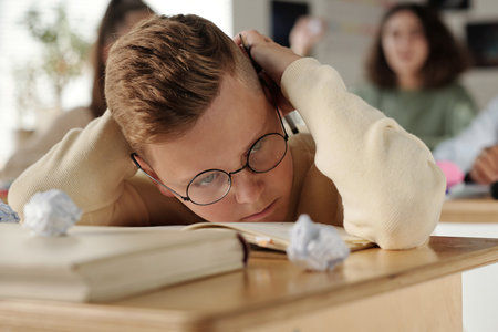Offended and upset schoolboy bending over desk to protect his head from crumpled papers thrown by classmates bullying himの写真素材