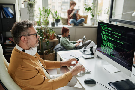 Confident mature businessman looking at coded data on computer screen while sitting by workplace in...