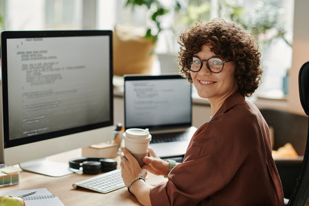 Young successful IT specialist with cup of coffee looking at camera while sitting by workplace with computer monitor and laptopの写真素材